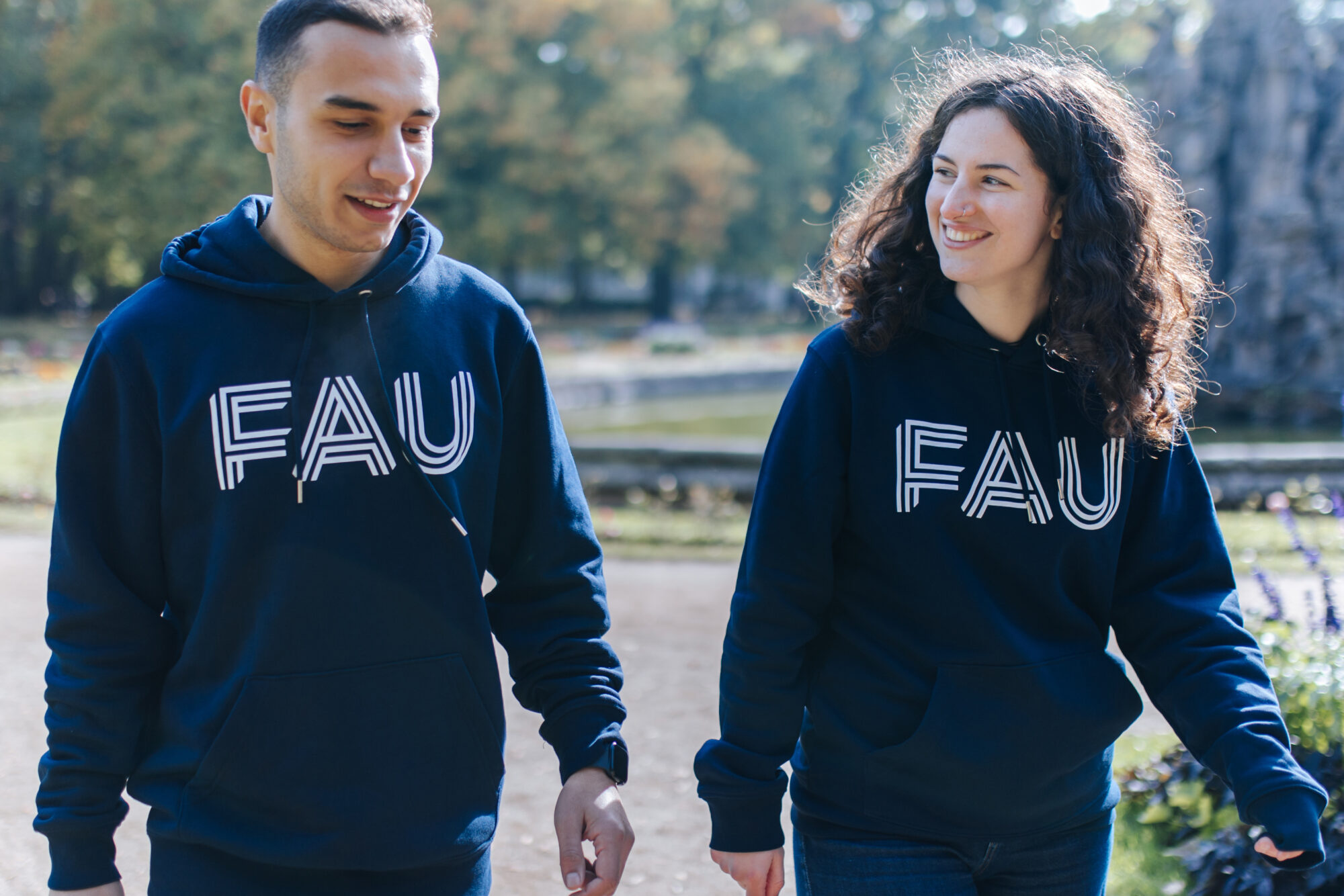 Ein männlicher und eine weibliche Student/in laufen gemeinsam durch einen Park und unterhalten sich. Sie haben beide FAU-Hoodies an.