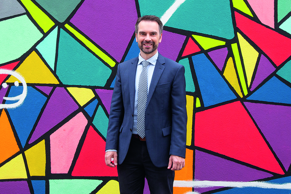 Ramming, ein Mann mit kurzen Haaren und Bart, steht vor einer bunten Wand.