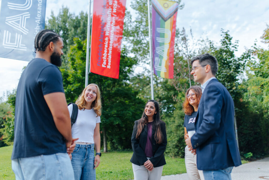 Eine GRuppe Menschen unterhält sich im Stehen. Im Hintergrund sind FAU-Fahnen zu sehen.