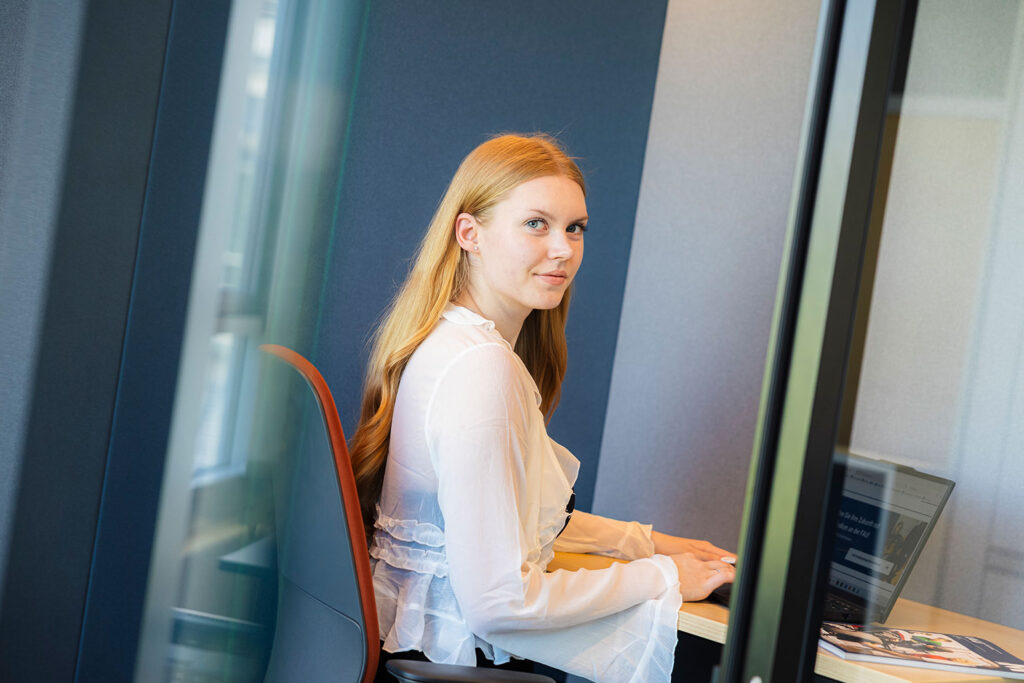 Eine Frau sitzt an einem Arbeitsplatz und arbeitet am Laptop. Sie schaut in die Kamera.