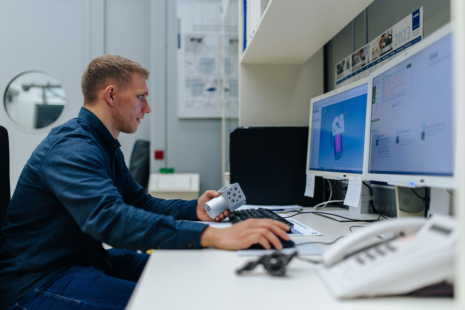 An FAU researcher sitting with a workpiece in front of two computer screens.