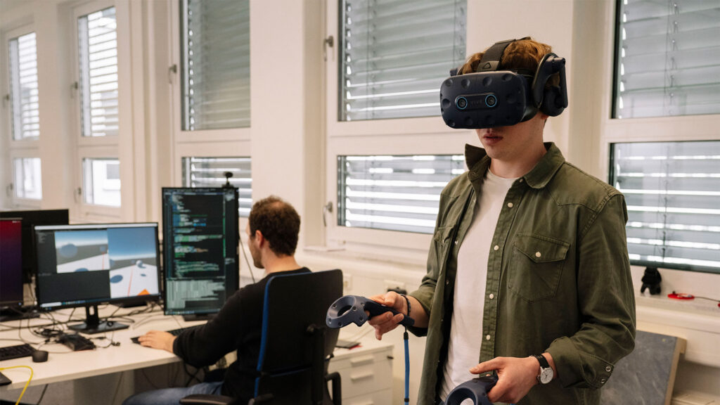 Im Vordergrund steht jemand mit einer VR-Brille auf dem Kopf, der mit zwei Controllern etwas ausprobiert. Im Hintergrund sitzt jemand vor Bildschirmen und überprüft den zugehörigen Code.