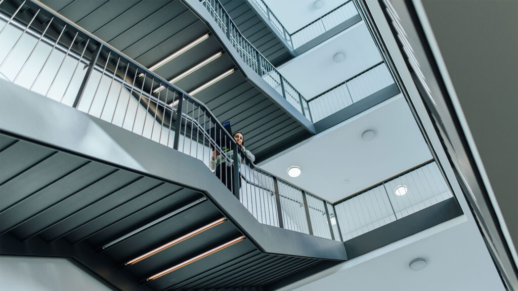 Ein helles Treppenhaus von unten. Man sieht mehrere Stockwerke. Eine junge Frau läuft mit einem Laptop abwärts und lächelt nach unten in die Kamera.