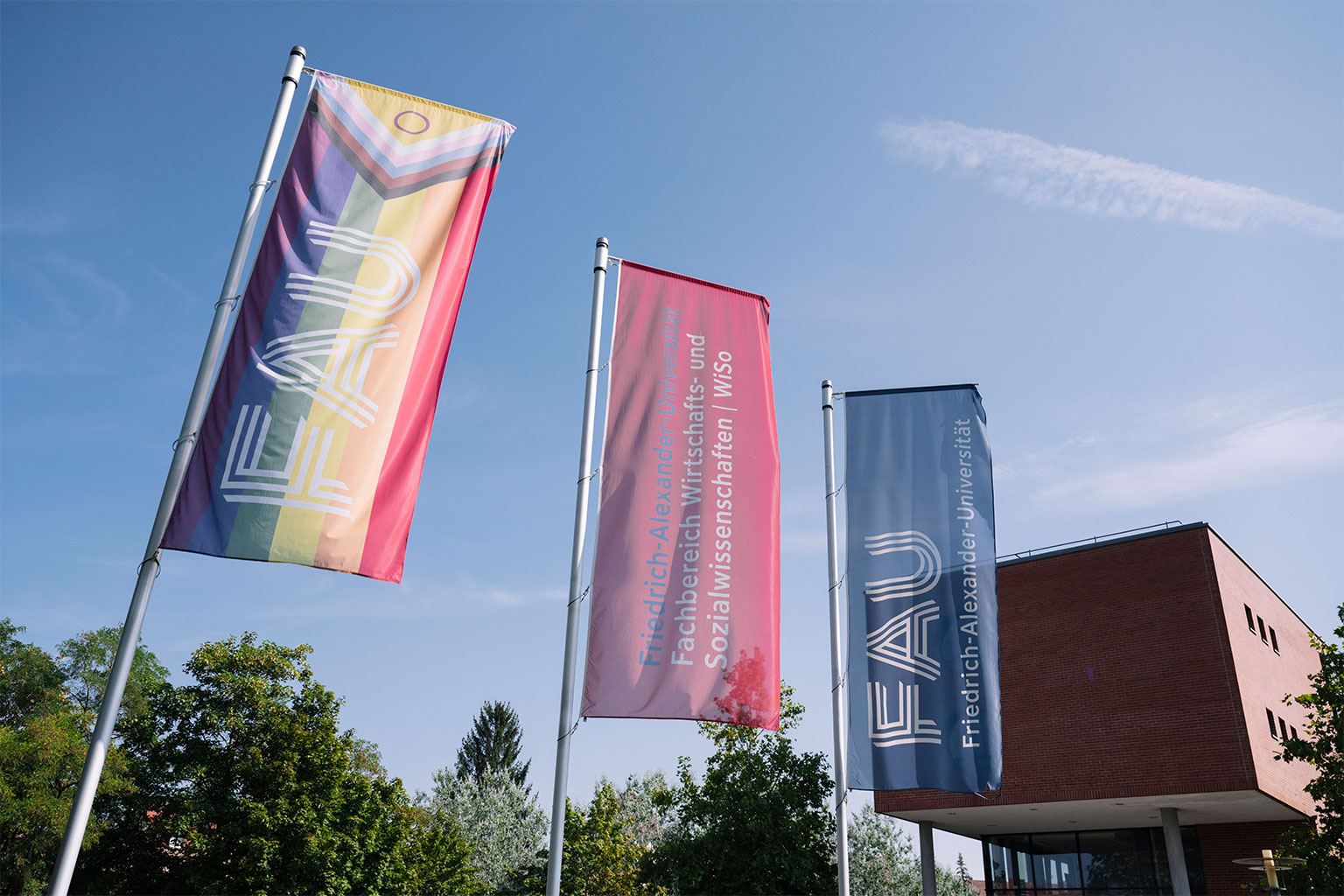 Es sind drei Flaggen mit dem FAU-Logo zu sehen. Die Flagge im Vordergrund ist die Regenbogenflagge. Im Hintergrund ist ein Teil eines Gebäudes der FAU zu sehen.